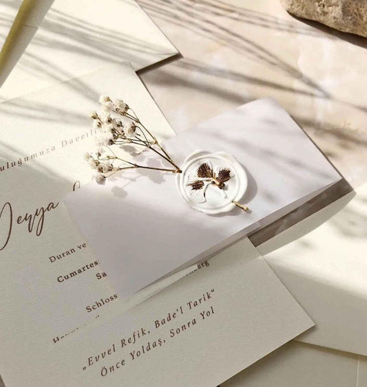 Elegante Hochzeitseinladung auf cremefarbenem Büttenpapier mit weißem Wachssiegel, Trockenblumen und feiner Schrift